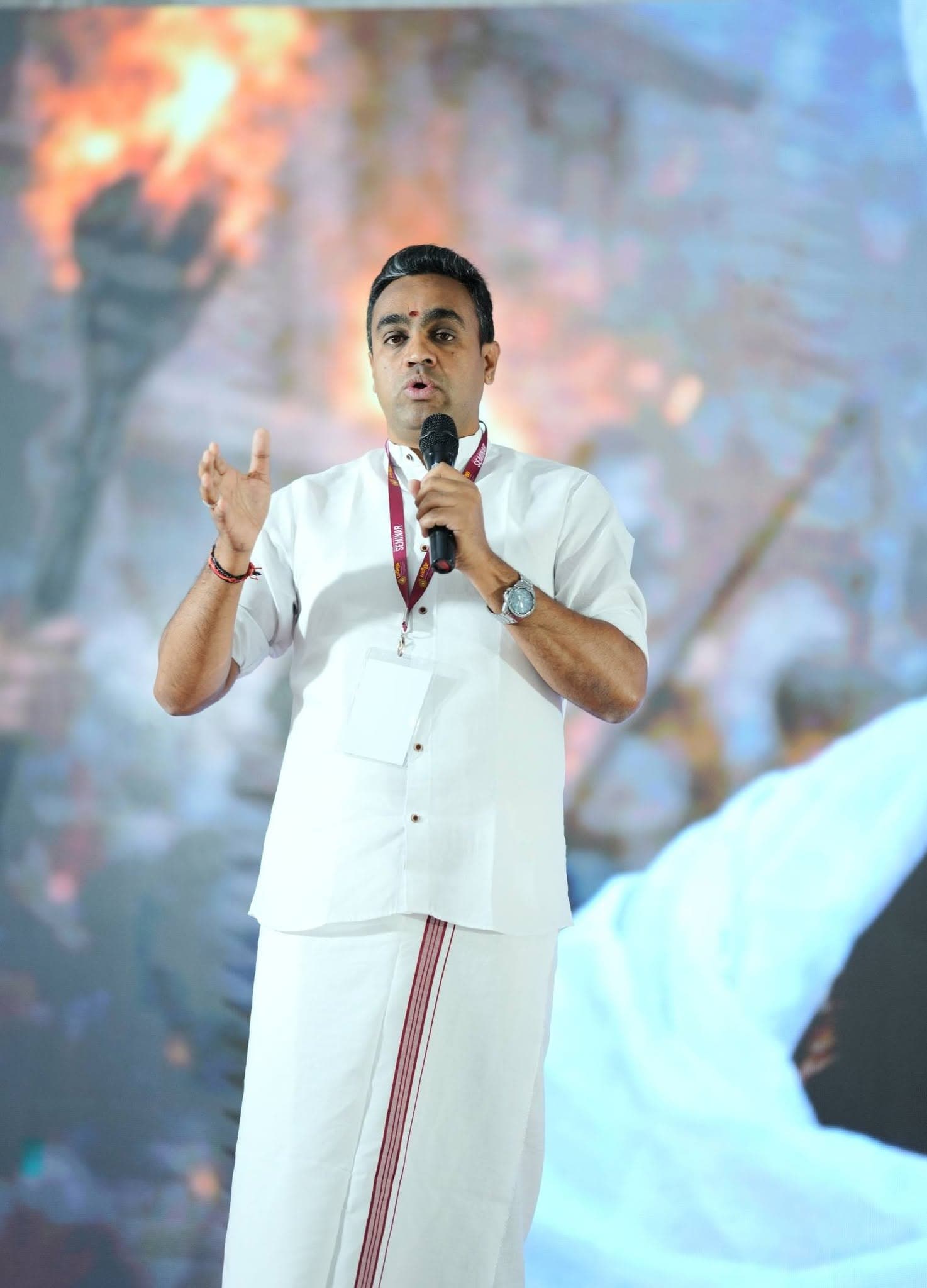 Manesh in traditional white veshti speaking with a microphone